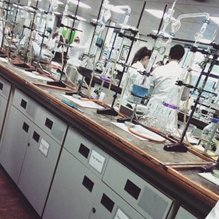 Inside the chemical analysis lab in the School of Food Science & Nutrition. Stands holding a variety of equipment including various flasks, burettes, & pipettes are set up on the work top. Two people in lab coats can be seen working in the background.