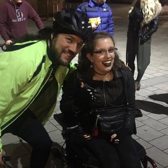 Wyatt from Planet Rock in high-vis cycling gear, including his helmet, leaning awkwardly in for a photograph. He had cycled 200 miles at this point. I'm sat in my wheelchair in my leather jacket, with black lipstick on.