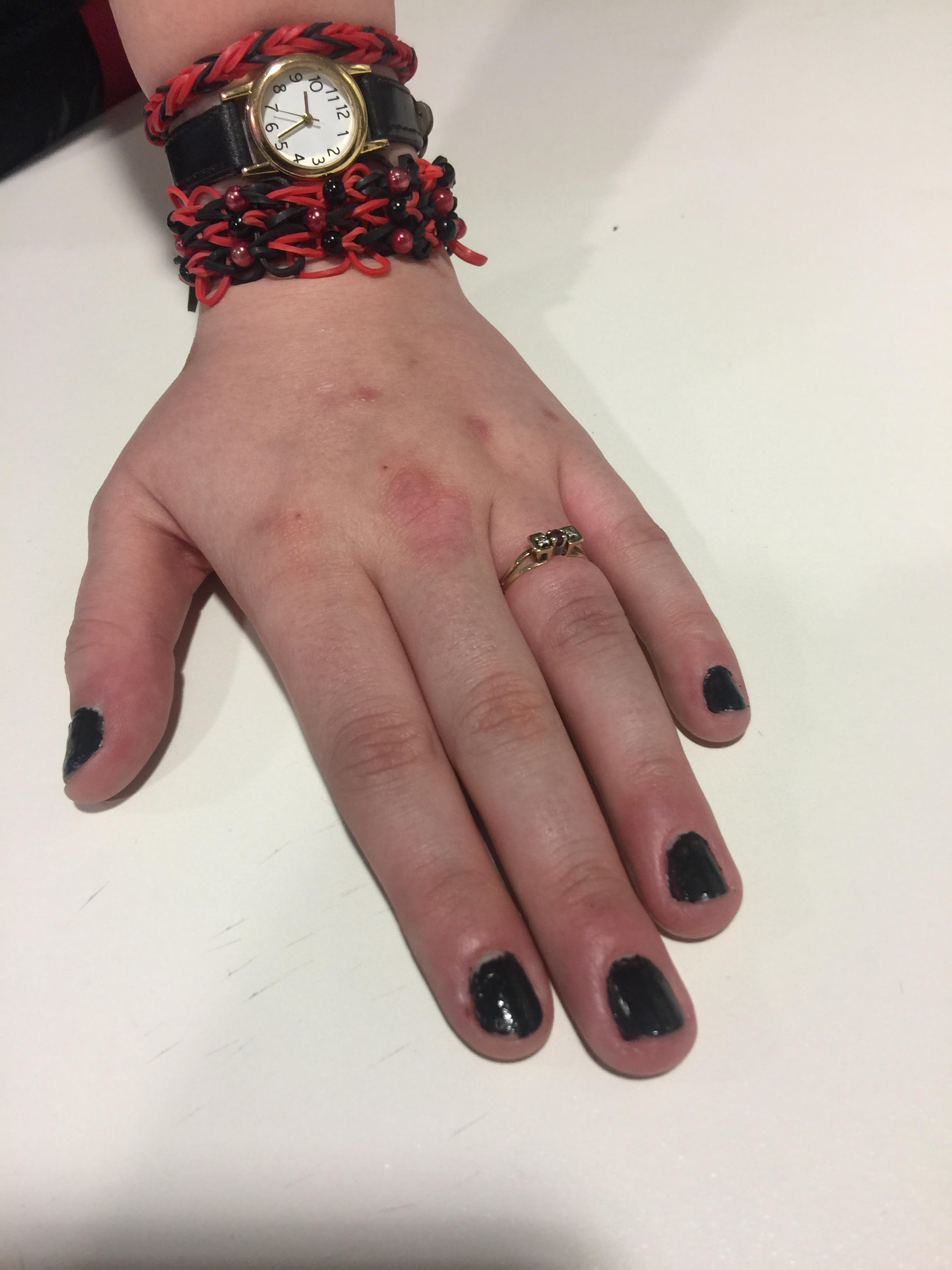 My left hand flat on the white table top, showing off my engagement ring. It's a simple gold band with 1 diamond in between 2 rubies on top. I have painted my nails black & am wearing a watch.