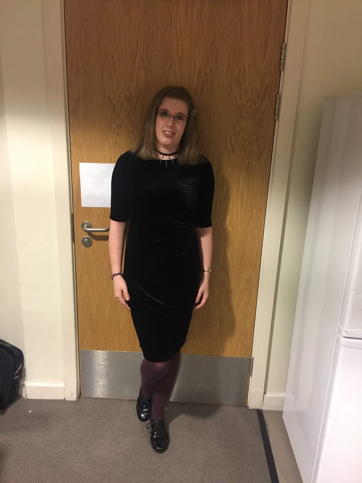 Stood up in my student residence. I'm wearing black heels, plum-coloured tights, & a plain black dress. The dress is made of a velveteen material and is form-fitting. My hair is straightened and loose.