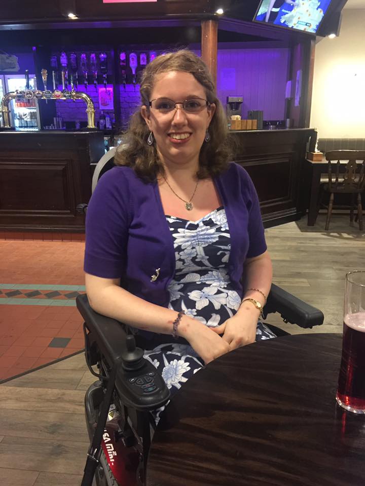 Image description: Sat in my powered wheelchair in a student bar. I'm wearing a blue dress with large flowers printed on, with a purple cardigan. My hair is half-up, half-down. This was taken on my 21st birthday.