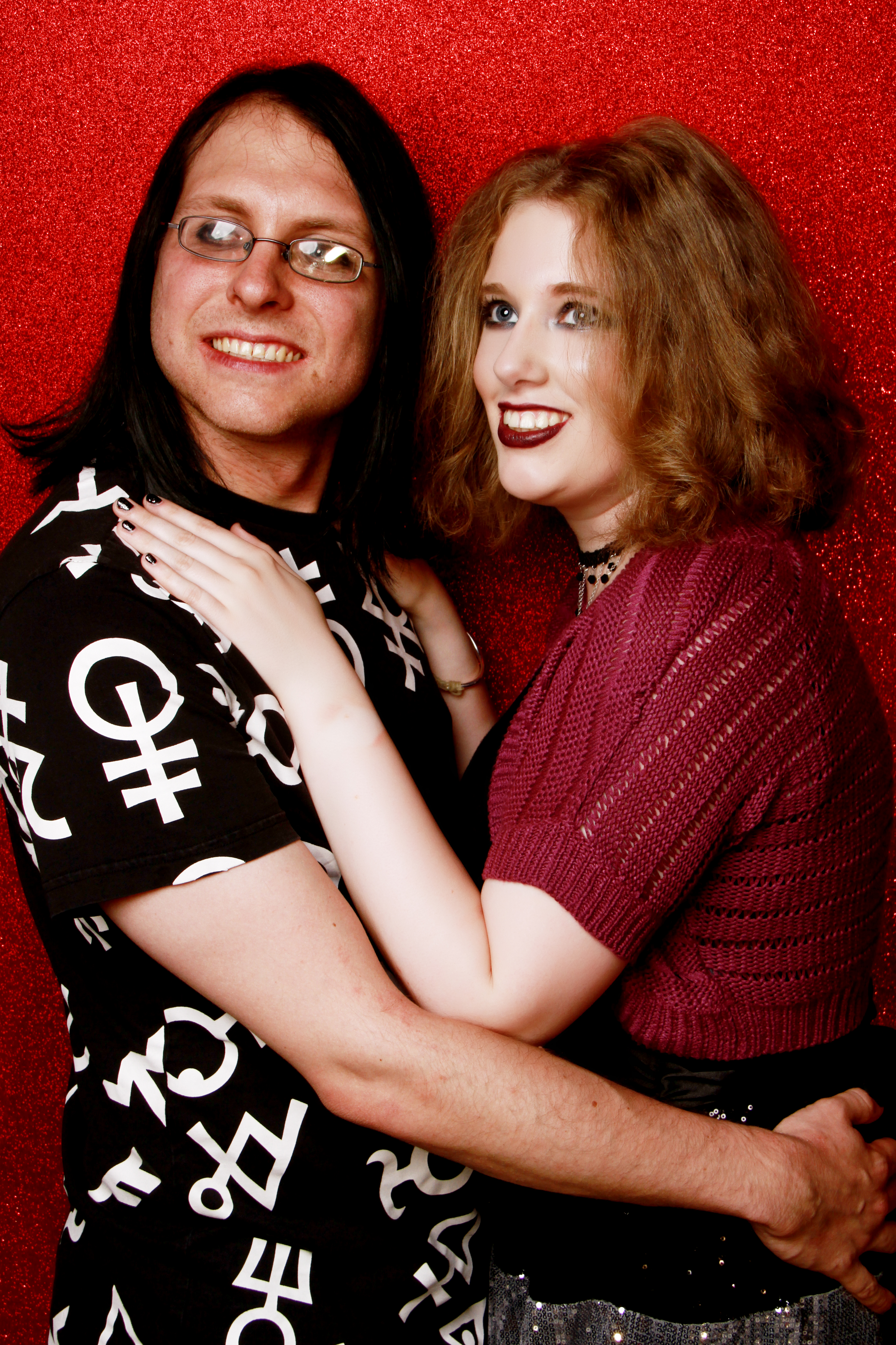 Image description: taken at a photo shoot. My husband and me are stood side-on to the camera, bodies facing each other but looking towards the camera. My hands are on his shoulders. I'm wearing my purple cardigan, & a black dress with a silver sequined skirt. Jarred is wearing a black t-shirt covered in occult symbols.