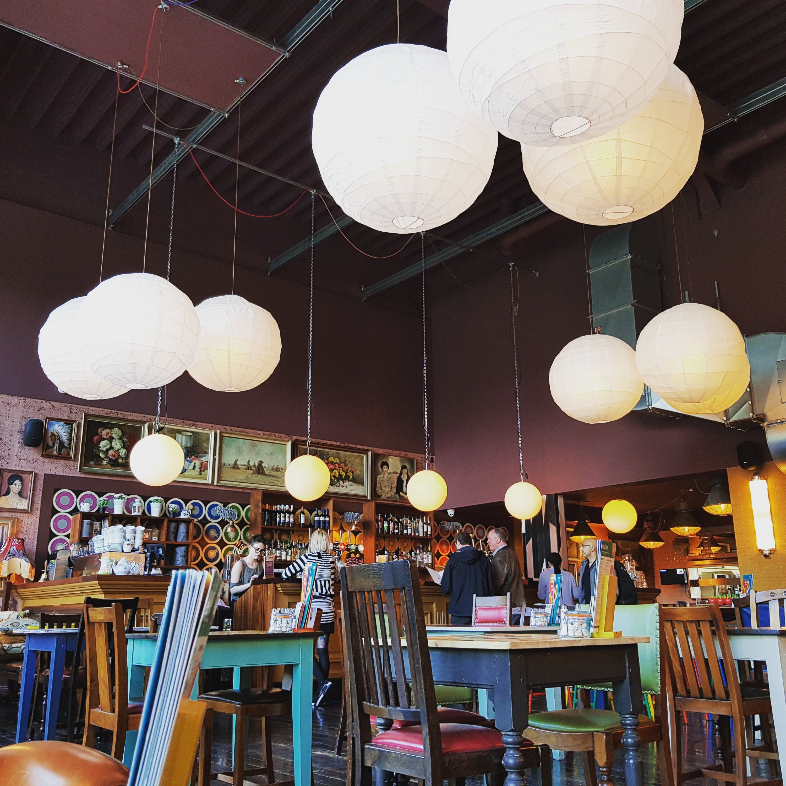Inside the cocktail bar. The maroon ceiling is high with cream, spherical lamp-shades dangling from it. The chairs are mismatched around similar tables, & there are a handful of people at the bar.