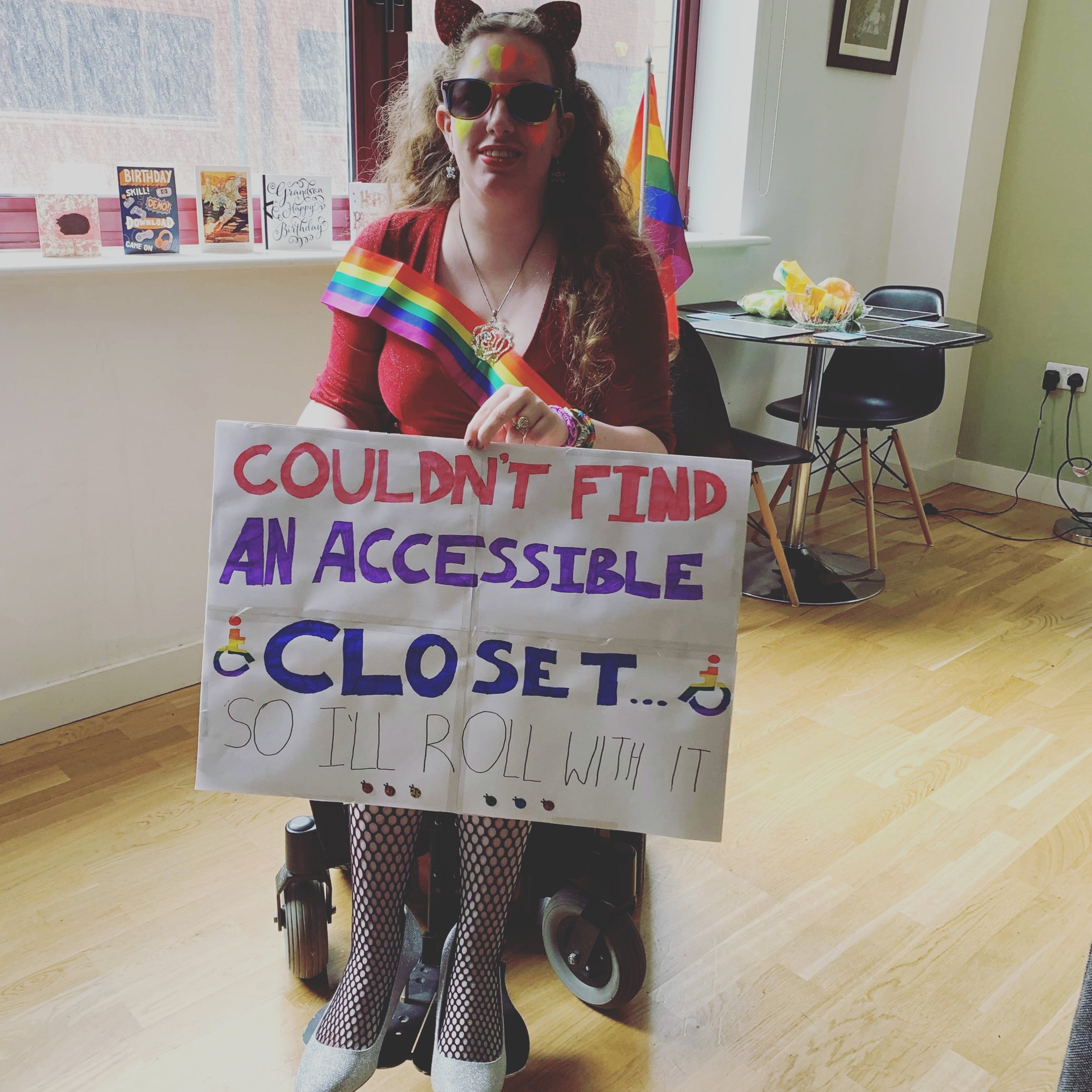 Sat in my wheelchair before pride, holding a handmade sign that reads "couldn't find an accessible closet so I'll roll with it." I'm wearing a red dress, fishnet tights, & silver heels.