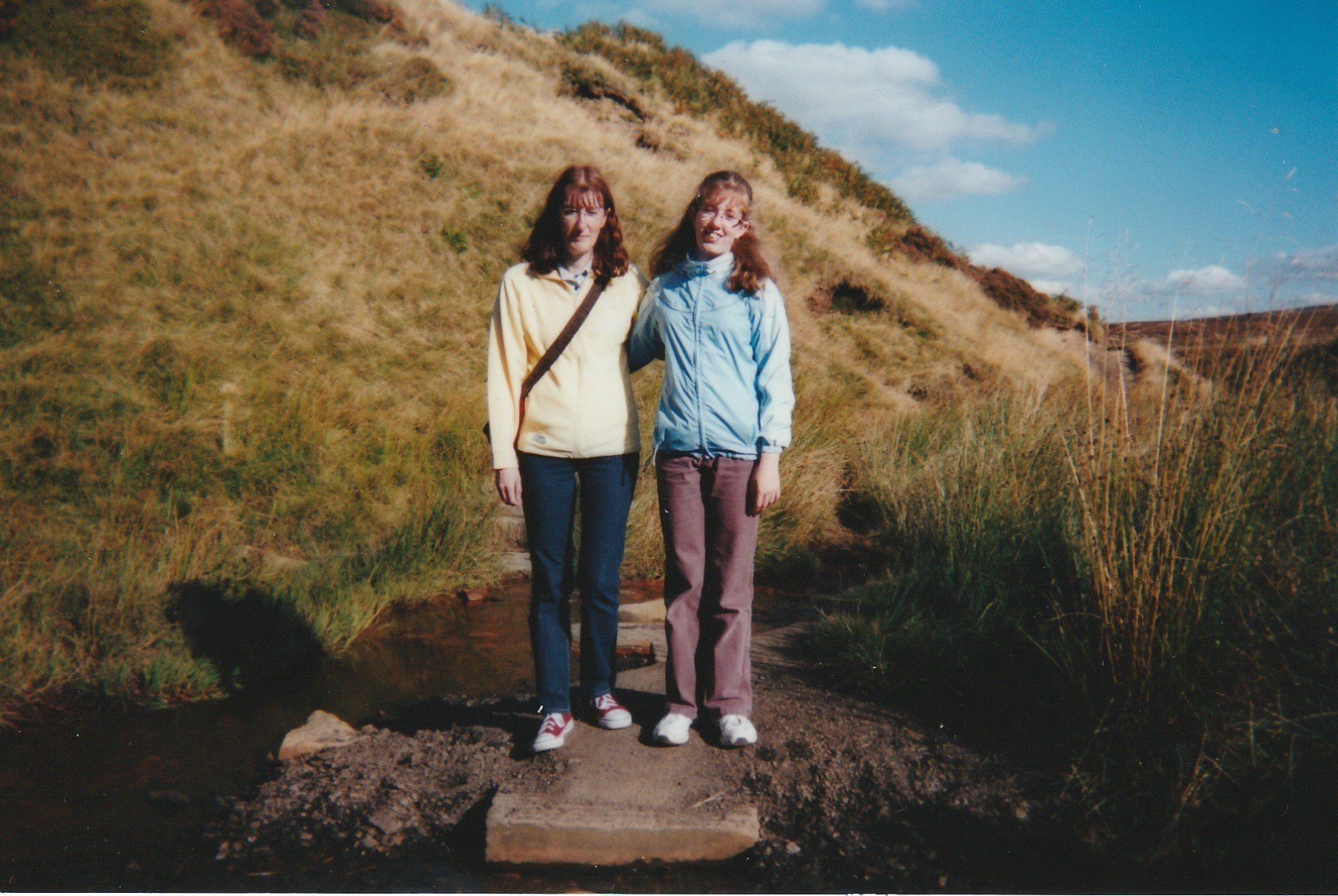 Stood with my mum, dressing in a yellow top & deep blue trousers, in the moors near Top Withins. We look very alike, with the same frizzy hair, & even tilting out heads the same way.