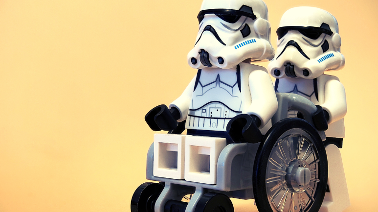 Two Lego Storm Troopers, one pushing the other in a wheelchair. Yellow backdrop.