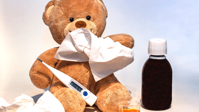 A teddy bear holding a tissue and thermometer sitting by a medicine bottle.