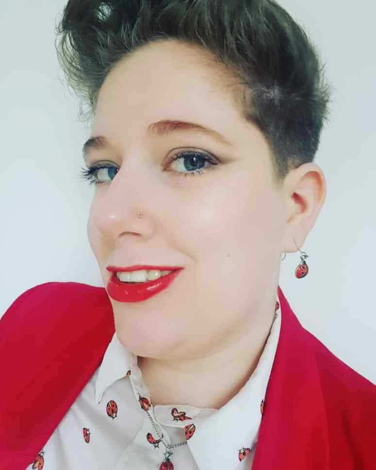 A selfie taken before my job interview. I'm sporting a red blazer over a white shirt dotted with ladybirds. I have ladybird earrings and a necklace on, and bright red lipstick.