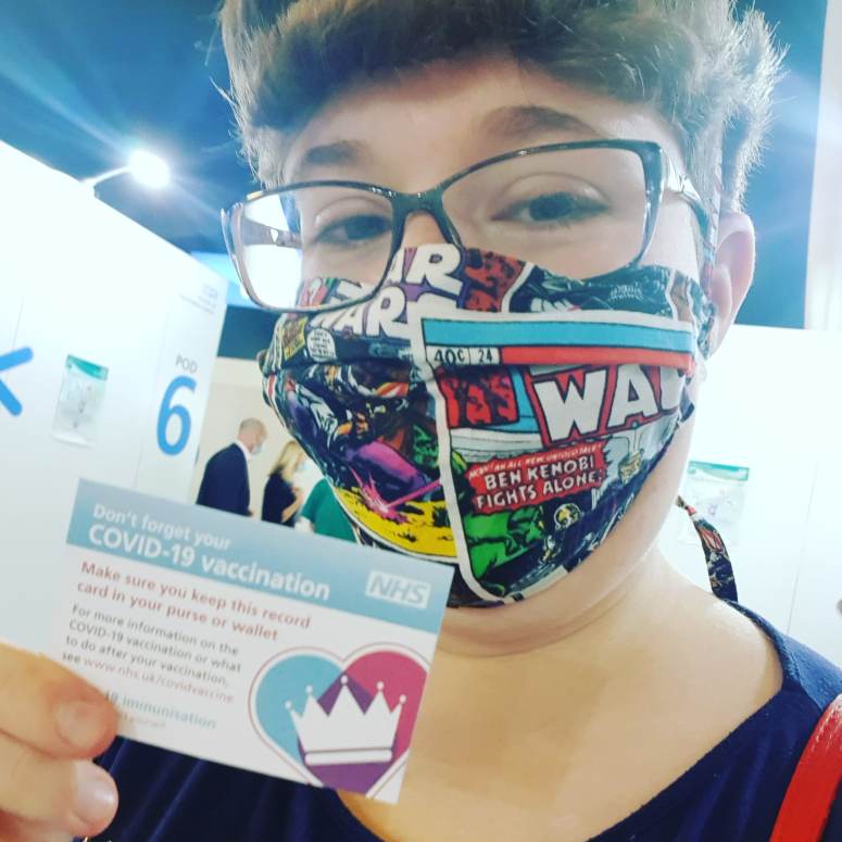Selfie taken in the vaccine centre, wearing a mask and holding up my proof of vaccination card.