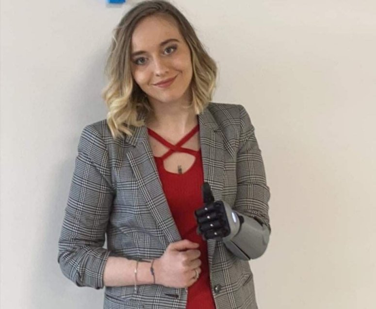 Wiki in a red top and grey jacket, doing a thumbs up with her bionic arm.