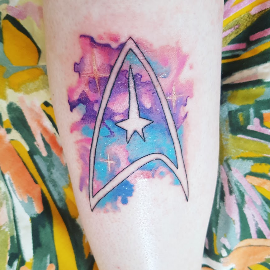 My freshly-done Star Trek logo tattoo filled with different shades of blue, pink and purple in a galaxy pattern.