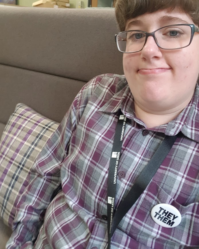 A selfie taken in my new work area, where my purple and grey checked shirt is almost identical to the cushion next to me. I look bemused.