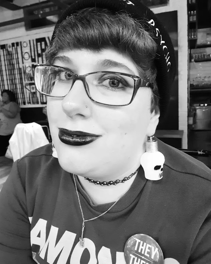 Black and white selfie taken in a bar. I'm wearing my Ramones sweater and a black beanie, with a tattoo choker, glowing skull earrings, and black lipstick.