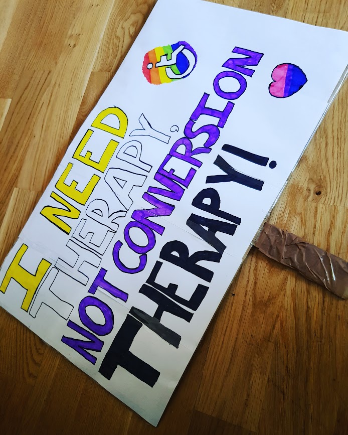 My pride sign reading "I need therapy, not conversion therapy" in capital letters. The text is displayed over four rows coloured yellow, white, purple and black like the non-binary flag.