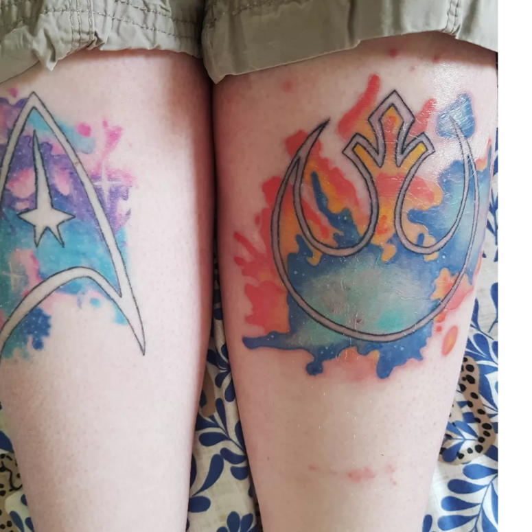 A close up of my legs showing both last years blue and pink Star Trek logo tattoo, and this year's orange and blue Star Wars rebellion logo. The Star Wars tattoo still has visible redness in places.