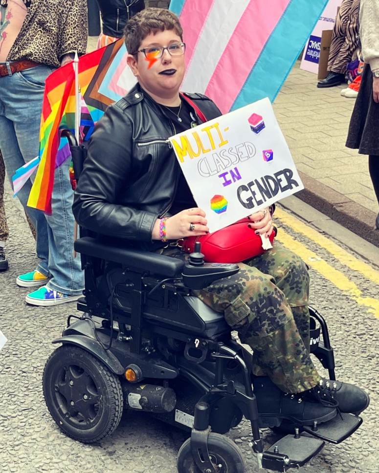 I sit in my powered wheelchair wearing camo pants and a black leather jacket, a David Bowie style lightning bolt painted on my cheek, surrounded by pride flags. I am hold a sign that reads Multi-classed in gender in the non-binary colours with pride flag dice.