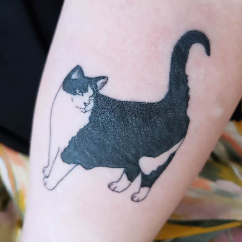 A close up of my arm showing the black and white tattoo of my beloved childhood cat, Bramble. She is stood facing left with her tail upright, and her head turned to look towards the right.
