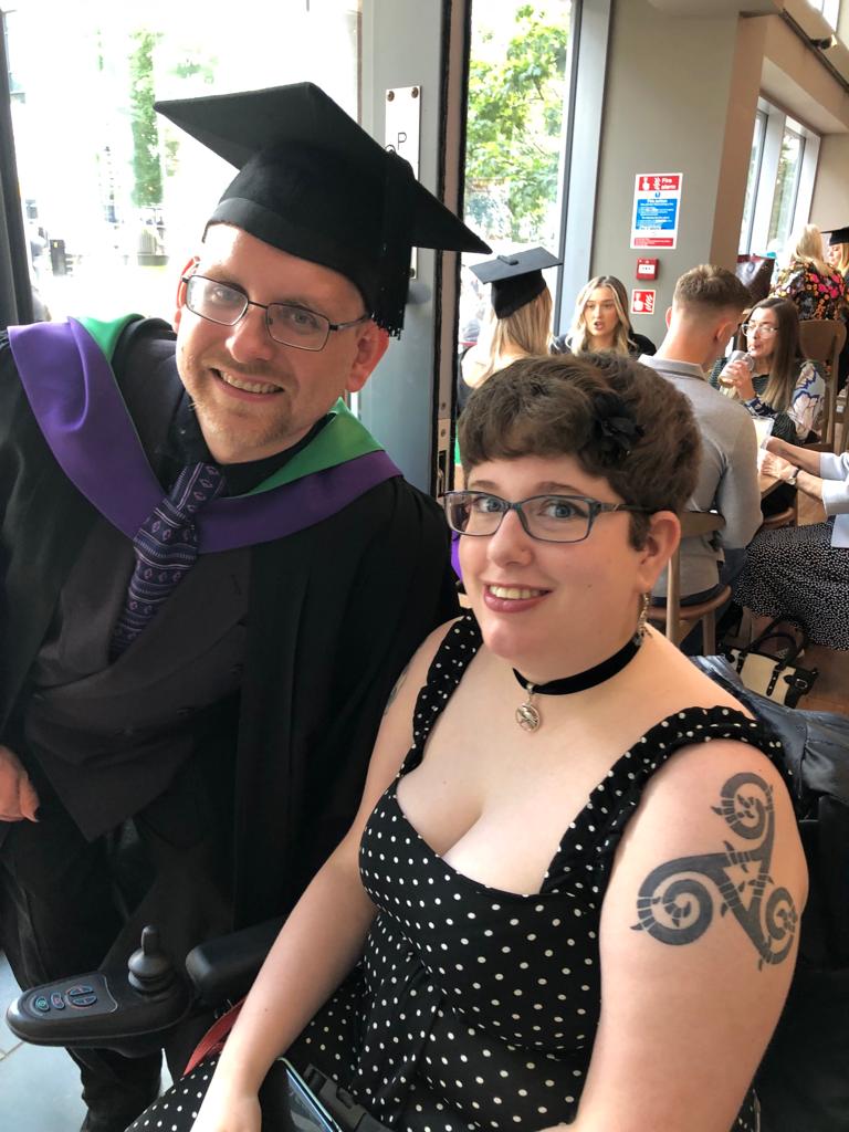 Me and my husband in a coffee shop ahead of his graduation. He is dressed in robes and cap, with a purple and green hood. I'm wearing a black pantsuit with white polka dots and a cute choker necklace, sat in my wheelchair. My triskele tattoo on my left shoulder is visible.