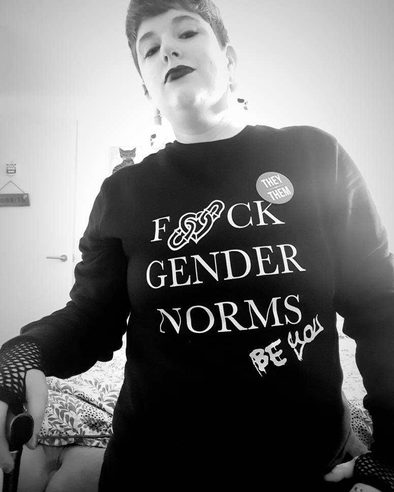 A black and white photo of me leant on the end of the bed, leaning on my walking stick with a fishnet gloved hand. I'm wearing a black sweater that reads Fuck Gender Norms, Be You, with a they/them pronoun badge pinned to it. I'm wearing dark eye makeup and black lipstick.